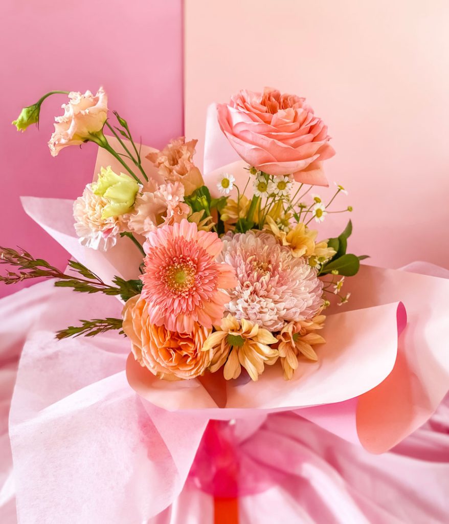 An arrangement of fresh flowers in pink, white, peach and yellow tones in a large glass mason jar wrapped with paper and ribbon