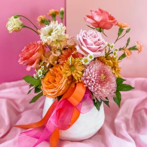 Large arrangement of pink, yellow, white and orange flowers in a white vase tied with ribbon