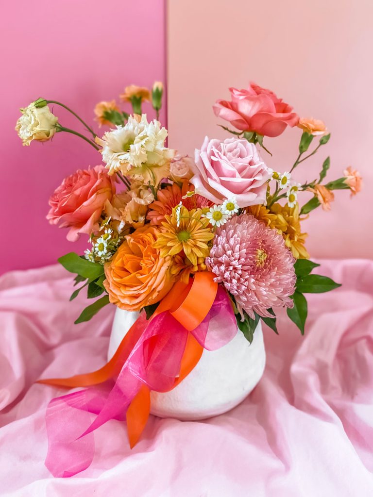 Large arrangement of pink, yellow, white and orange flowers in a white vase tied with ribbon