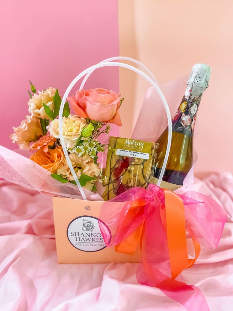 A gift bag tied with ribbon and filled with a bunch of fresh flowers in pink, white and peach tones along with a chocolate bar and bottle of wine