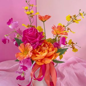 Arrangement of brightly coloured silk artificial flowers in pink, orange and yellow tones in a white textured vase tied with ribbon