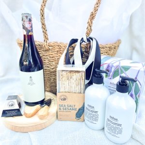 The image shows a bottle of wine, gourmet treats, and hand wash outside the basket.